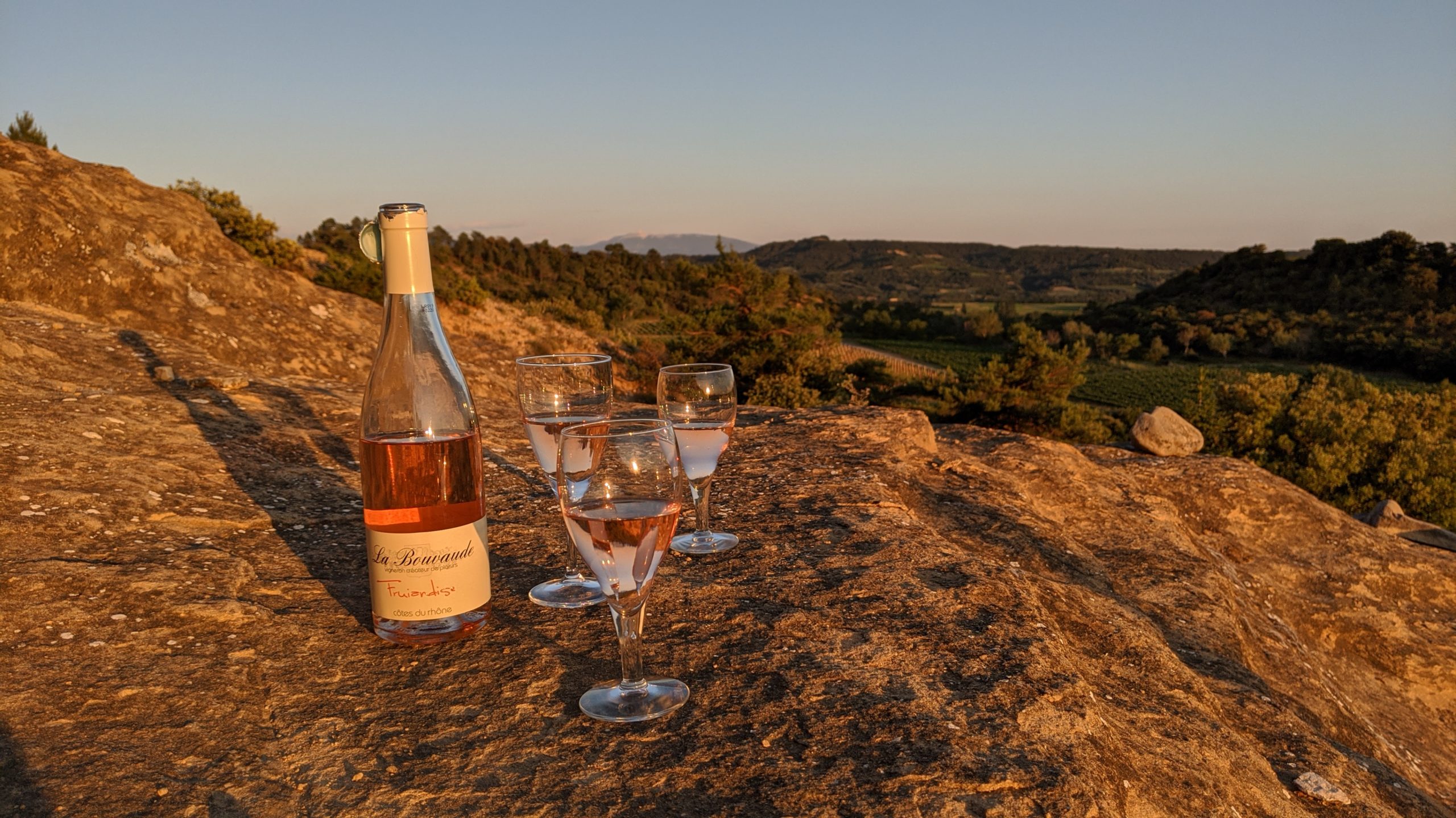 Une bouteille de rosé et trois verres de vin posés sur un rocher au coucher de soleil